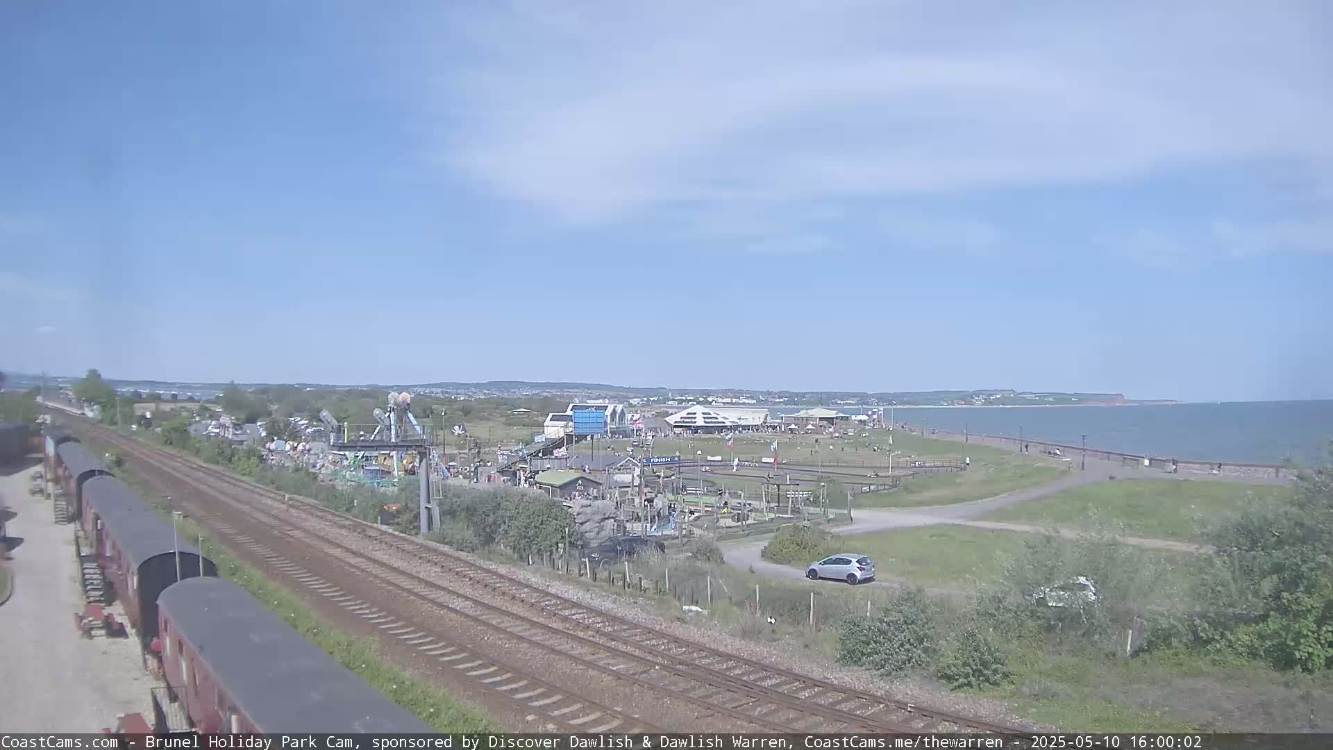 A seaside amusement park with a beachfront promenade is visible under a clear blue sky, with train tracks running alongside.