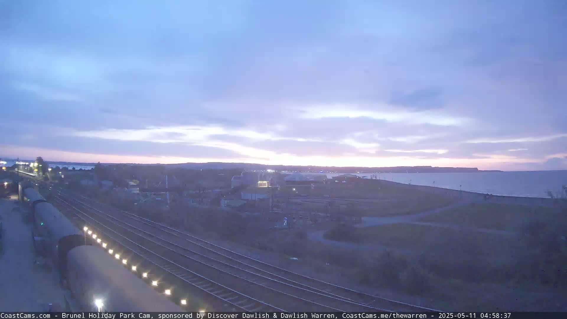 A twilight view shows train tracks near a coastline, with a calm body of water under a mostly clear sky.