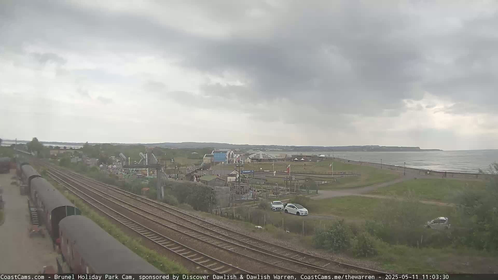 Overcast skies loom over a coastal amusement park and train tracks, with a calm sea visible in the distance.