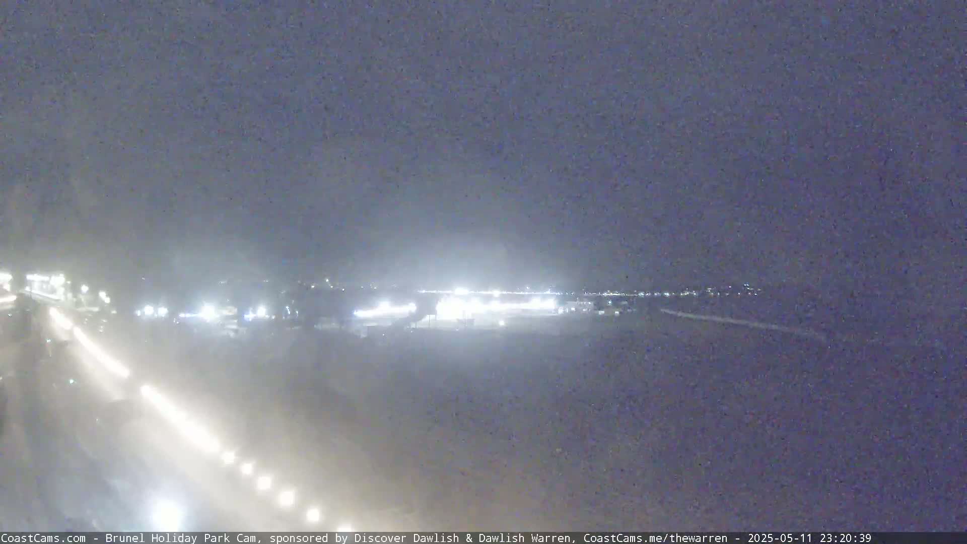 A nighttime view of a road with headlights visible in foggy or hazy conditions, showing distant lights of a town or buildings in the background.