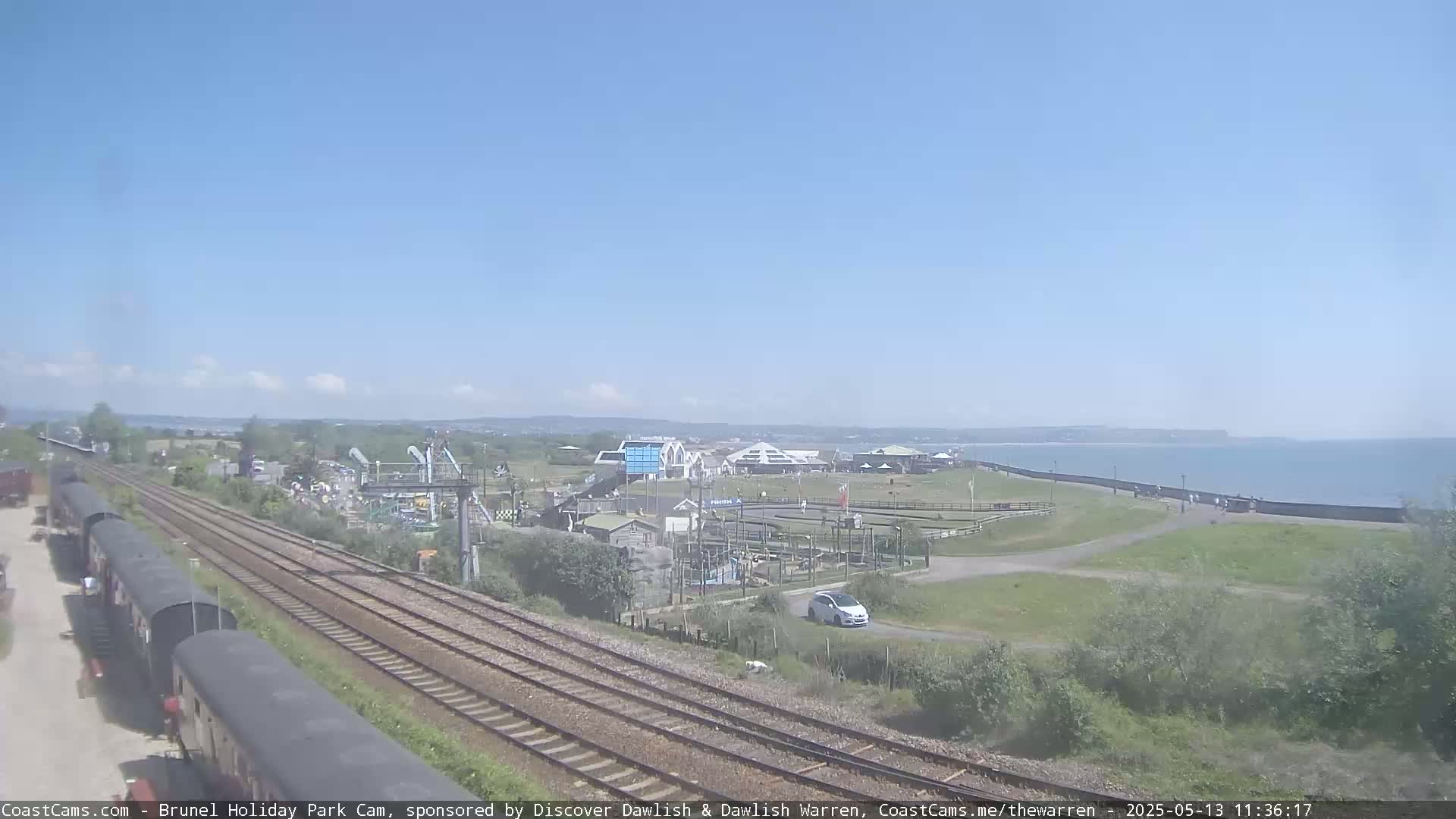 Under a clear blue sky, a coastal scene shows railway tracks next to a seaside park with a car, buildings, and a promenade alongside the ocean.
