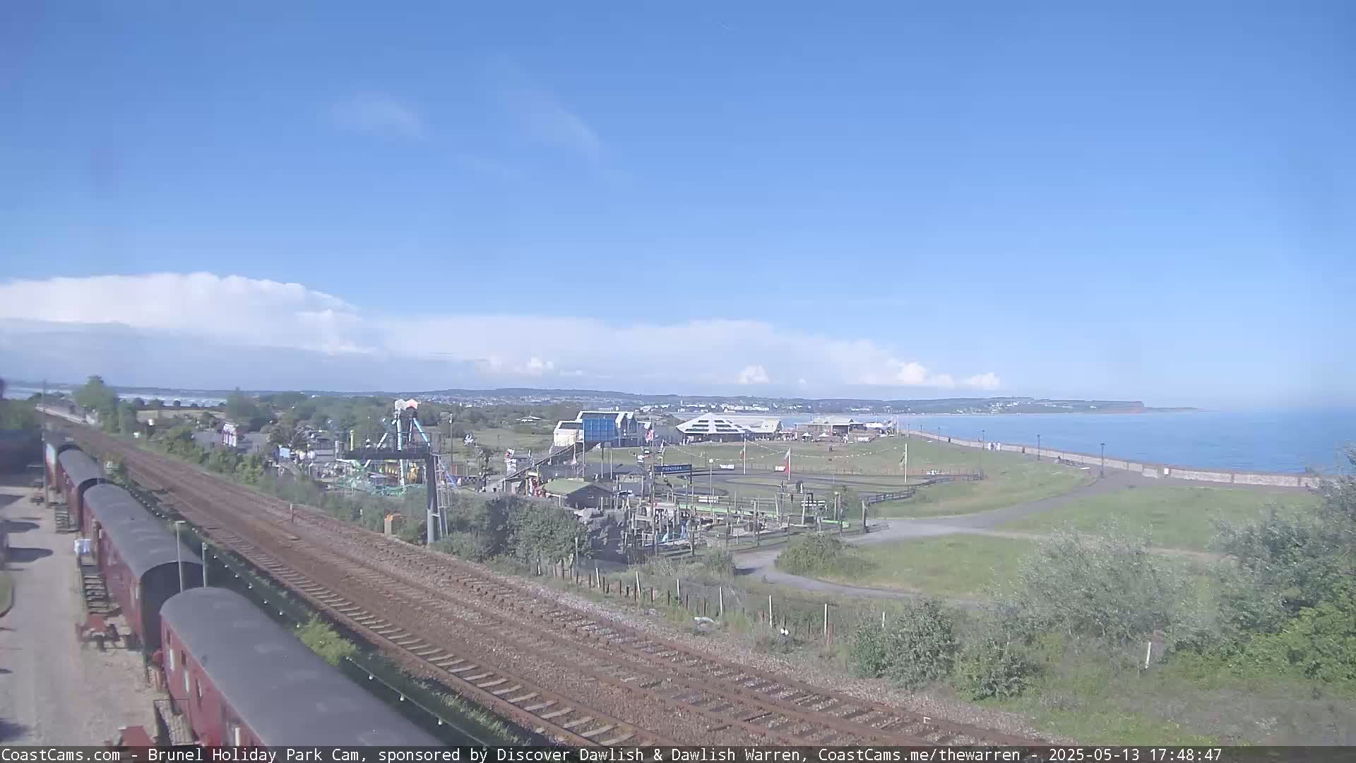 A coastal amusement park with a train track and several railway cars is visible under partly cloudy skies.