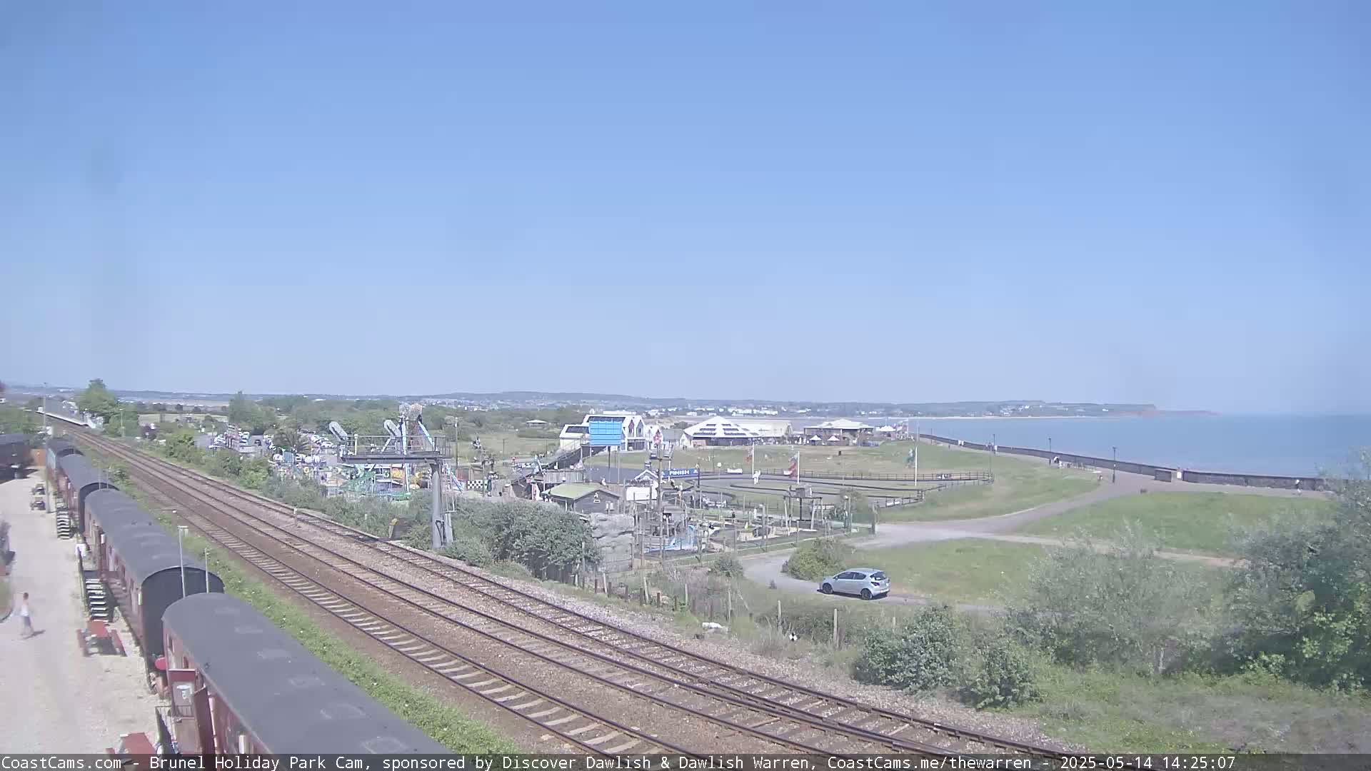 A sunny day reveals a coastal scene with train tracks running alongside a seaside amusement park and a calm ocean.