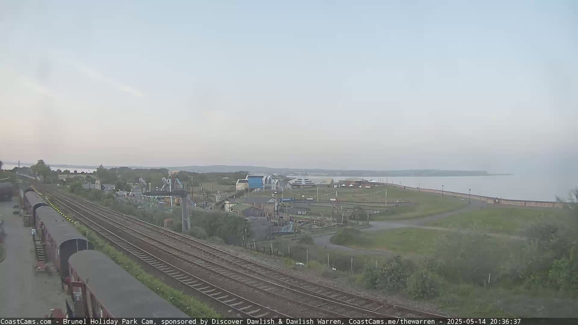 A railway line runs through a coastal park next to a calm body of water under an overcast sky.