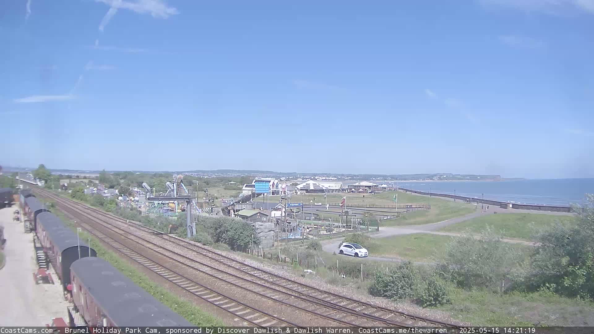Under a clear blue sky, a seaside amusement park and train tracks are visible, with a calm ocean in the background.