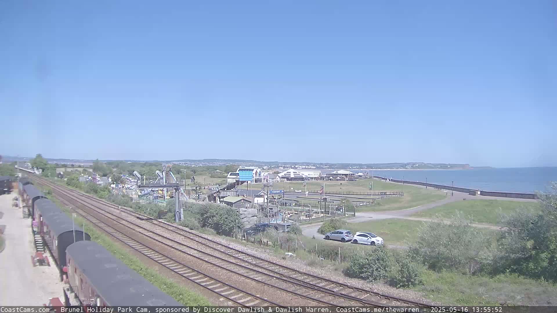A seaside amusement park with a go-kart track is visible near a railway line on a clear, sunny day.