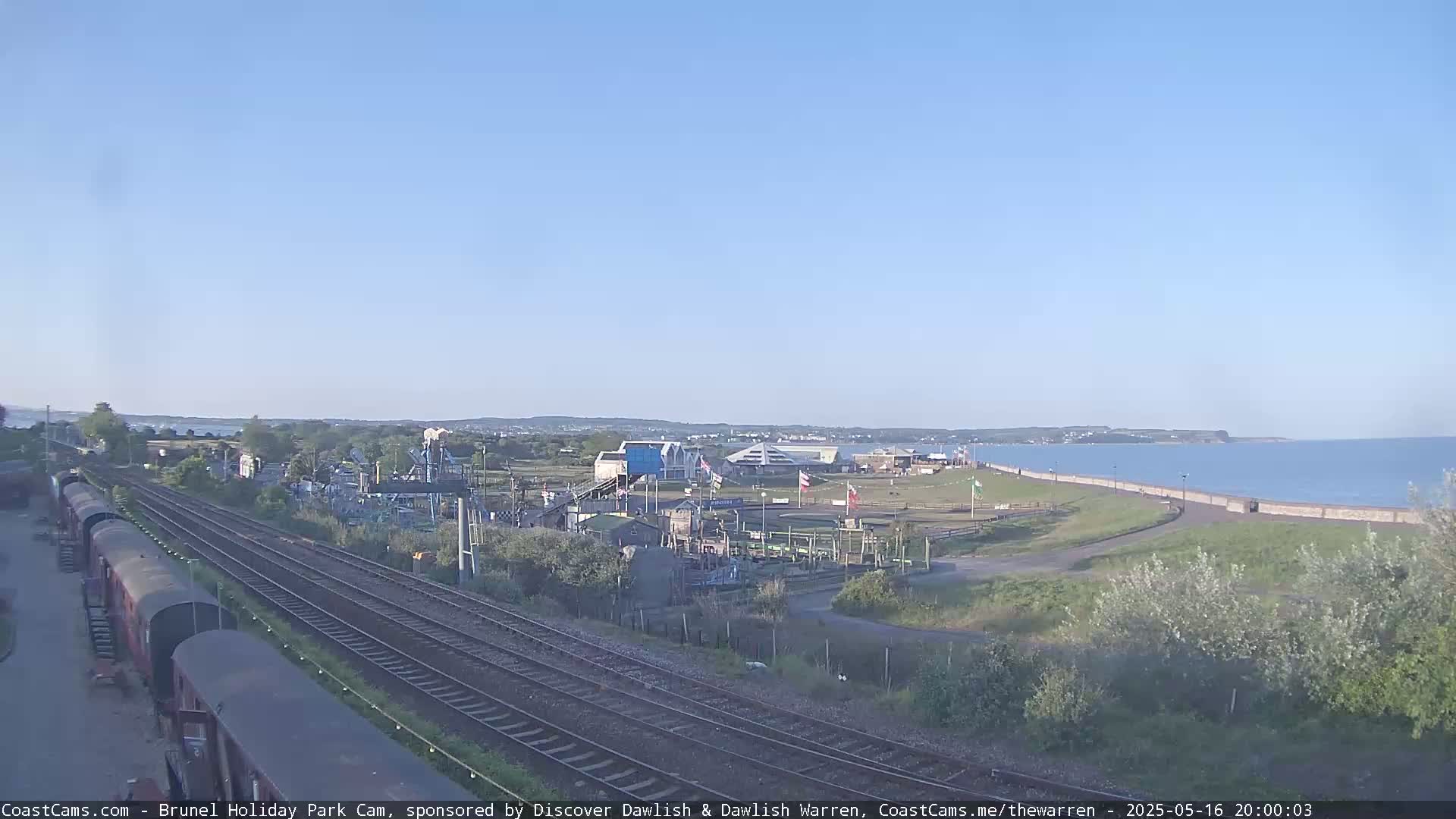 A clear, sunny day shows a coastal scene with train tracks running alongside a holiday park near a calm ocean.