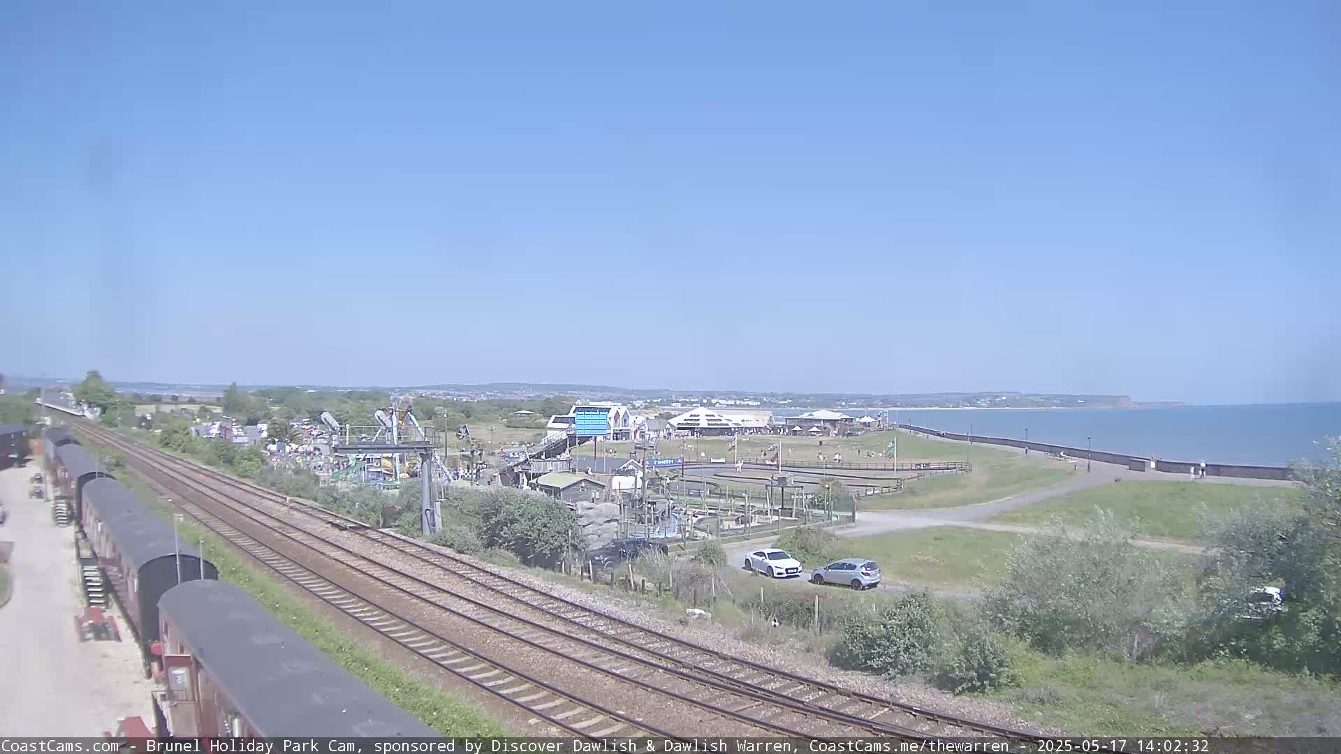 Under a clear blue sky, railway tracks run alongside a seaside amusement park toward a calm ocean.