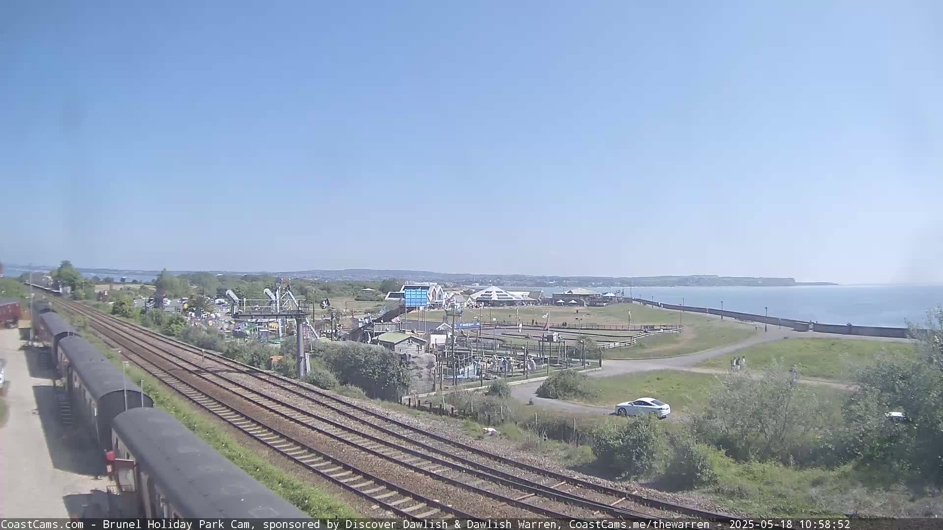 Under a clear blue sky, a coastal amusement park is situated beside a body of water, with railway tracks and several train cars visible in the foreground.