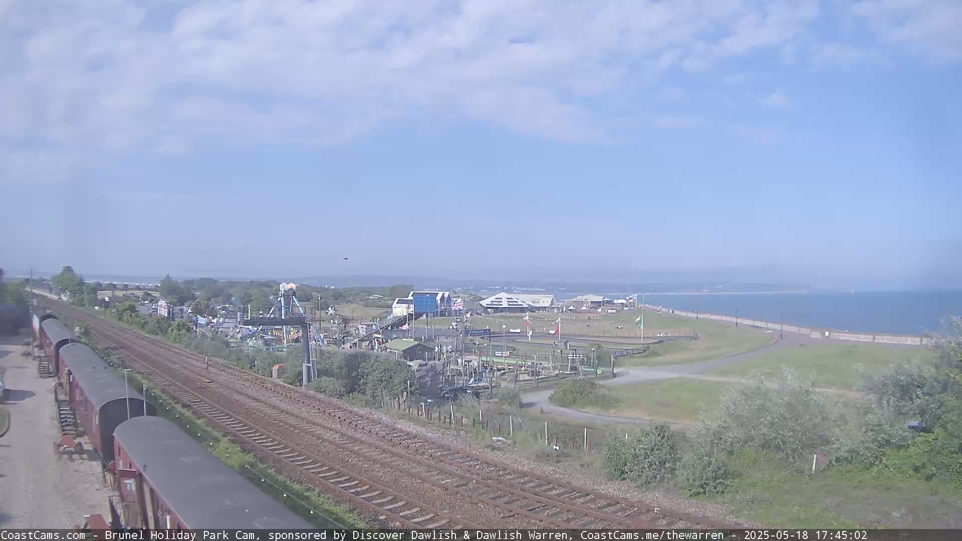 A seaside amusement park with train tracks beside it is visible under a mostly clear sky, with a calm body of water in the background.