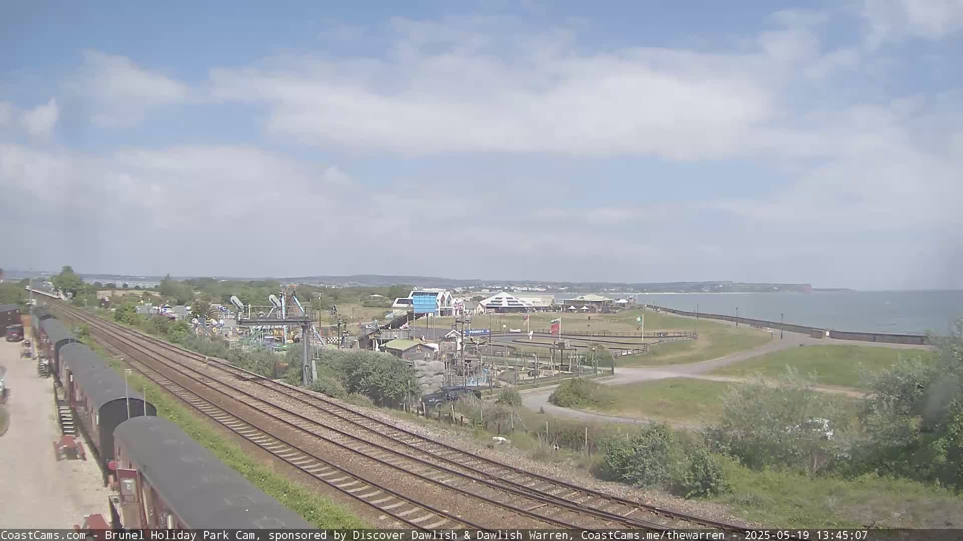 A seaside amusement park with a train track running alongside it is visible under a partly cloudy sky, with a calm ocean in the background.