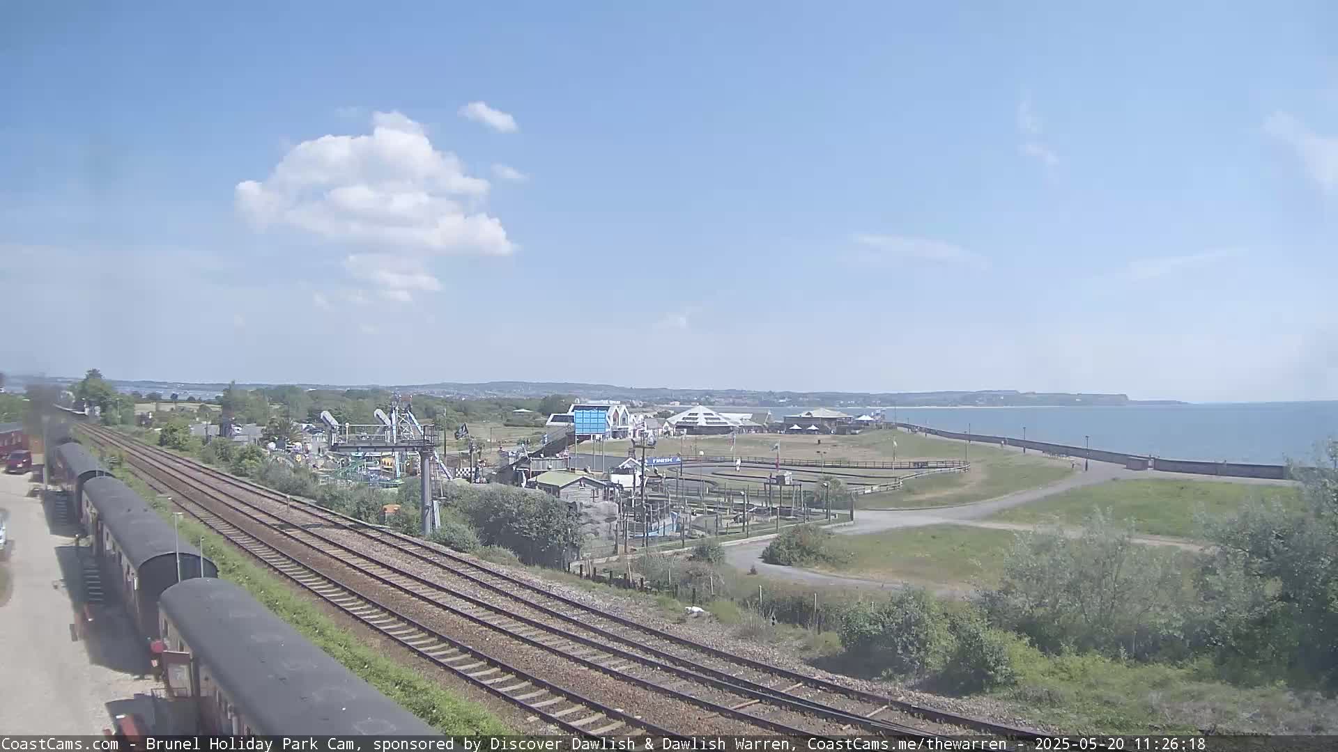 A sunny day reveals a seaside amusement park near train tracks, with a calm body of water in the background.