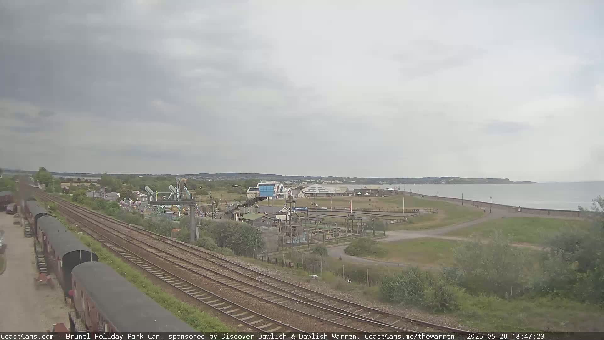 A seaside amusement park is seen from a distance, with overcast skies, railway tracks in the foreground, and a calm sea in the background.