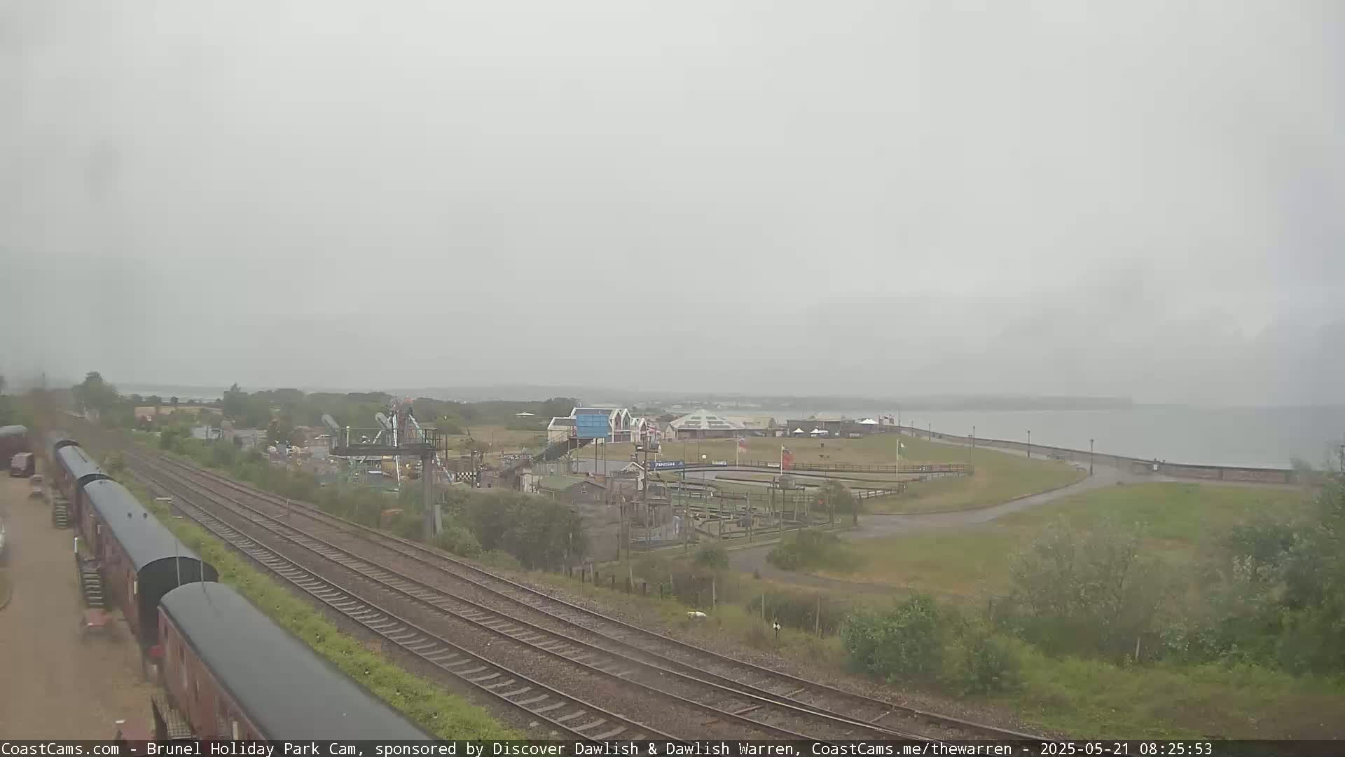 A hazy, overcast day reveals a seaside amusement park with train tracks running alongside it, leading to a body of water.