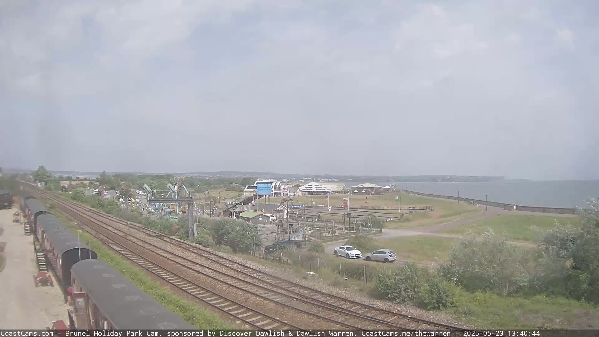 A seaside amusement park is visible next to railway tracks on a mostly cloudy day, with a calm body of water in the background.