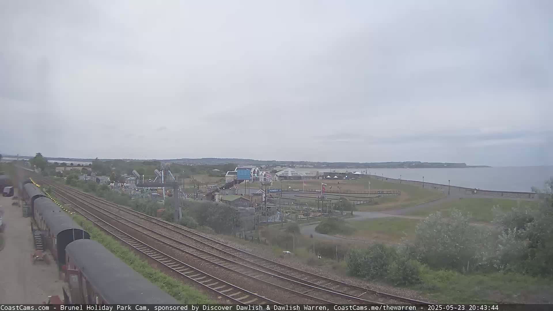 A seaside amusement park with a go-kart track is visible beyond railway tracks, under an overcast sky.
