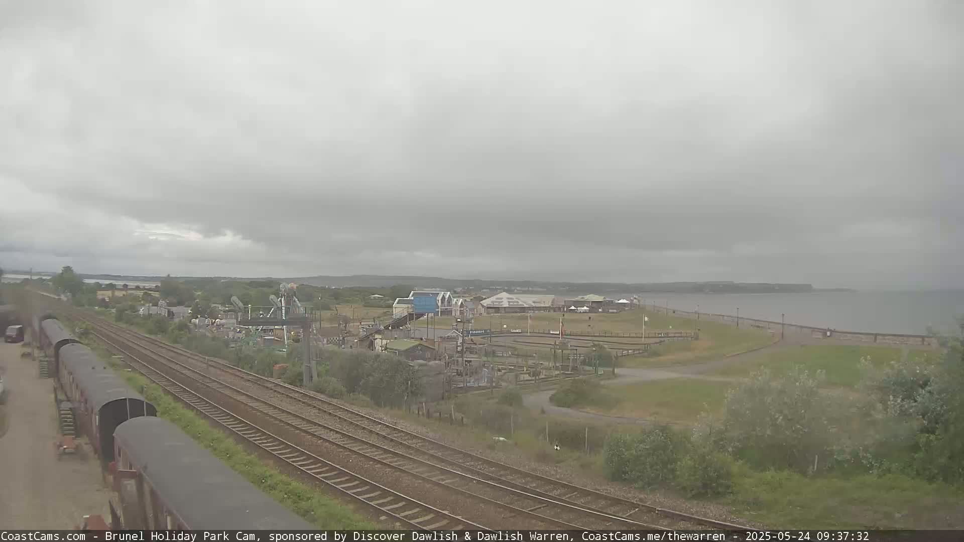 A seaside amusement park is visible beyond railway tracks under an overcast sky.