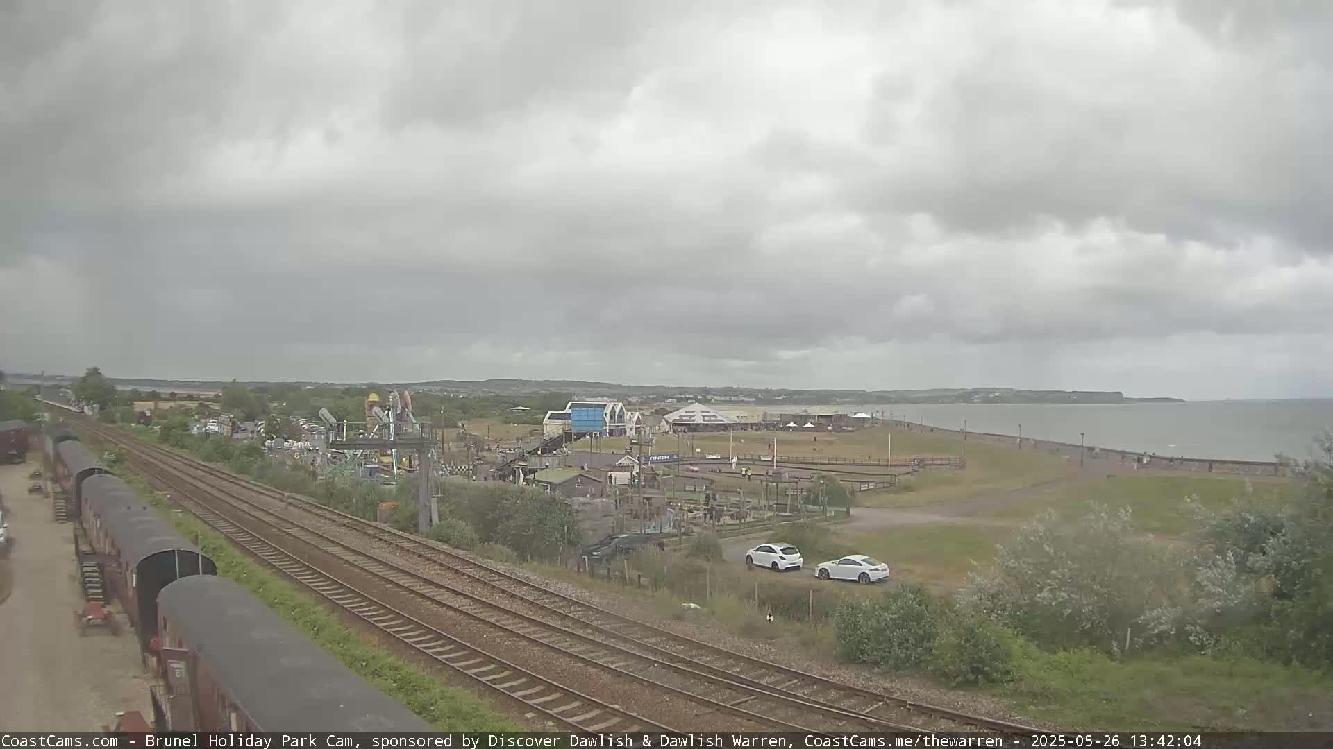 A seaside amusement park with overcast skies, a body of water, railway tracks, and several parked cars is visible from a high vantage point.