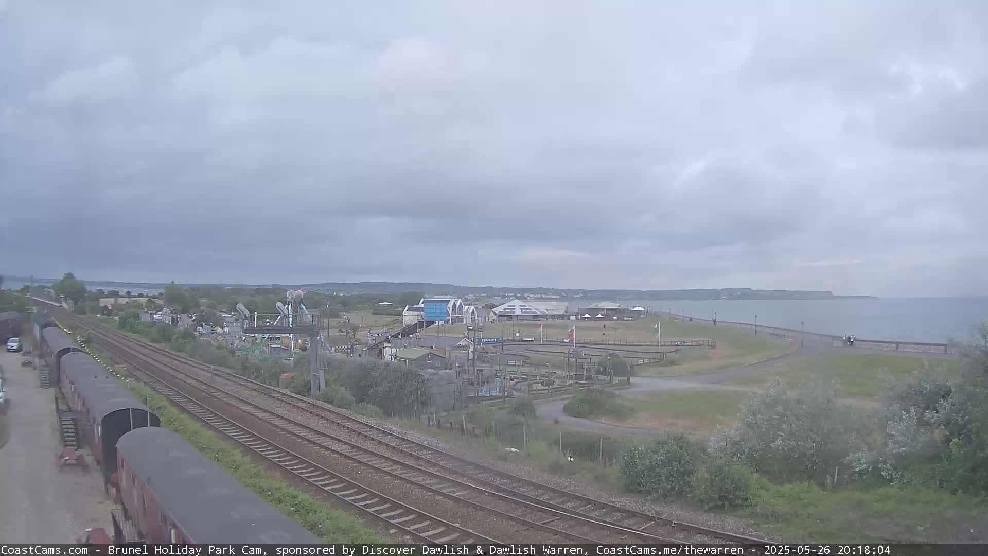 Under an overcast sky, a coastal amusement park with go-karts and other rides sits beside train tracks and a sea wall with a calm body of water beyond.