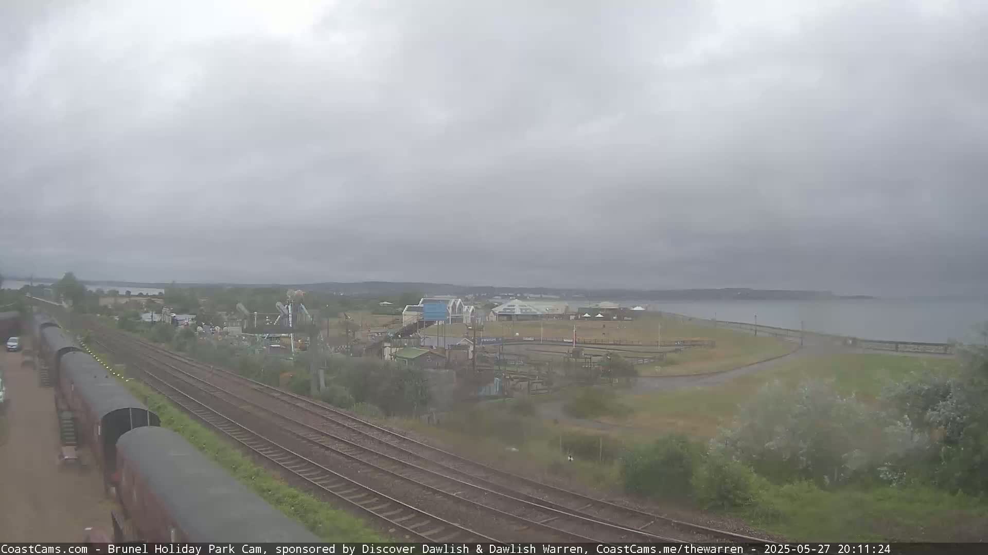 A high-angle, long shot shows a coastal area on an overcast day, with railroad tracks running through the foreground near a small amusement park and body of water beyond.