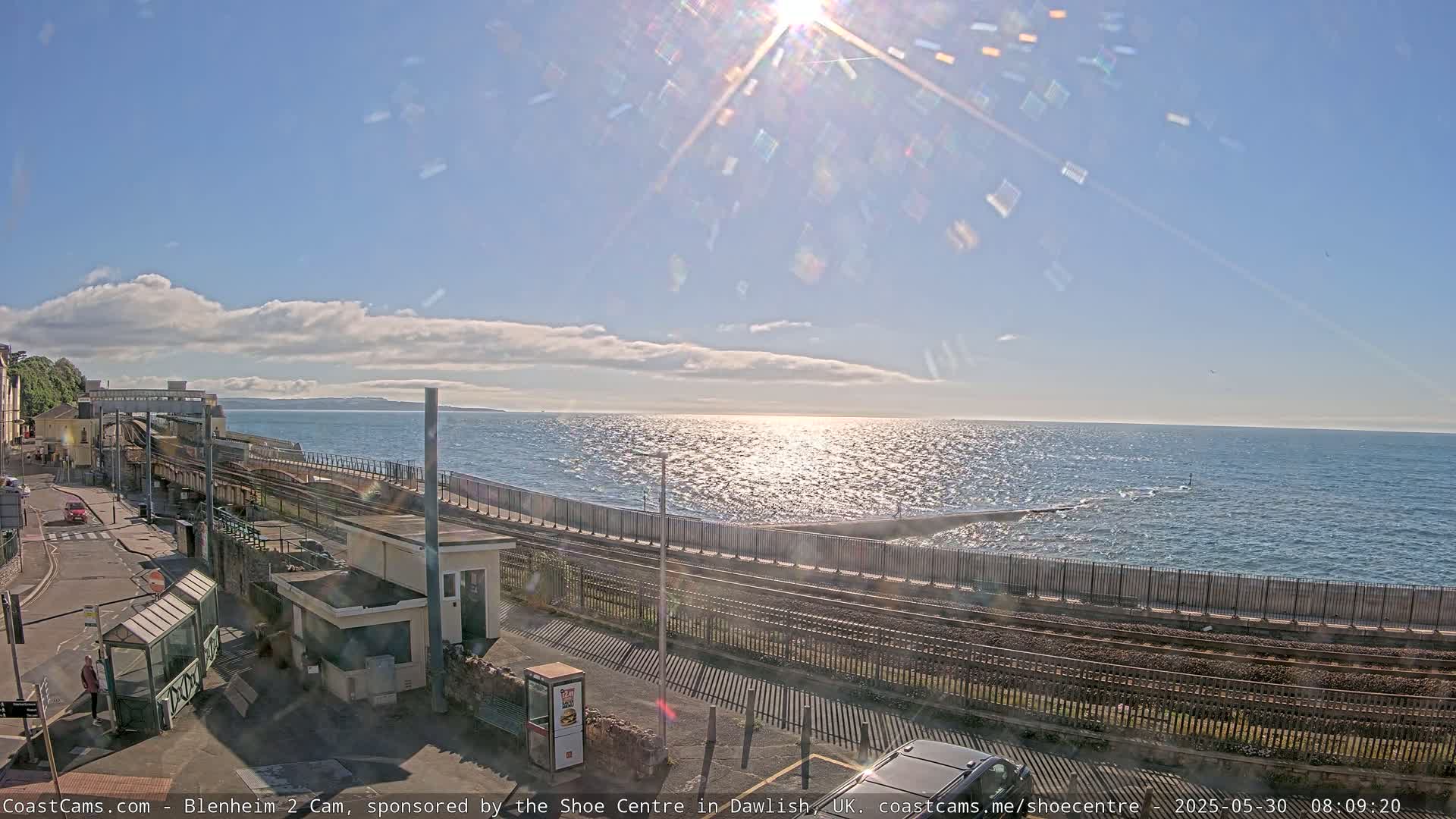 A sunny day shows a coastal train track next to a calm sea, with a building and a road visible.