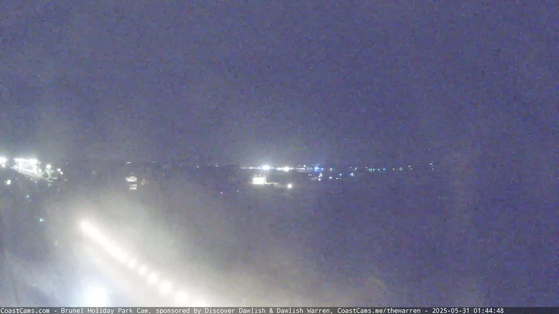 A nighttime long shot of a hazy, dimly lit landscape with a few lights visible in the distance.