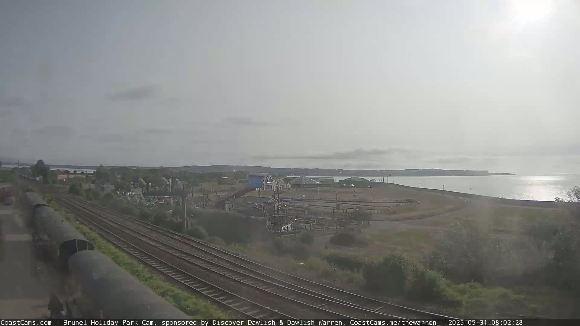 A coastal scene under an overcast sky shows a train track, amusement park rides, and a calm sea with a distant shoreline.