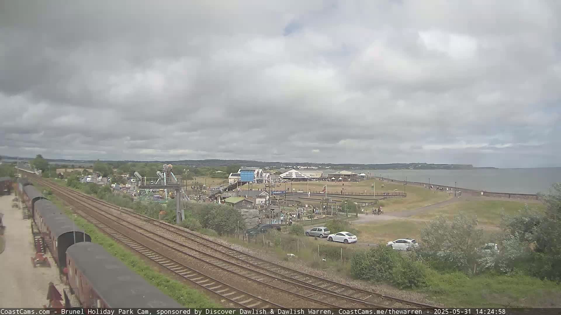 A seaside amusement park with overcast skies, railway tracks, and a calm body of water in the background.