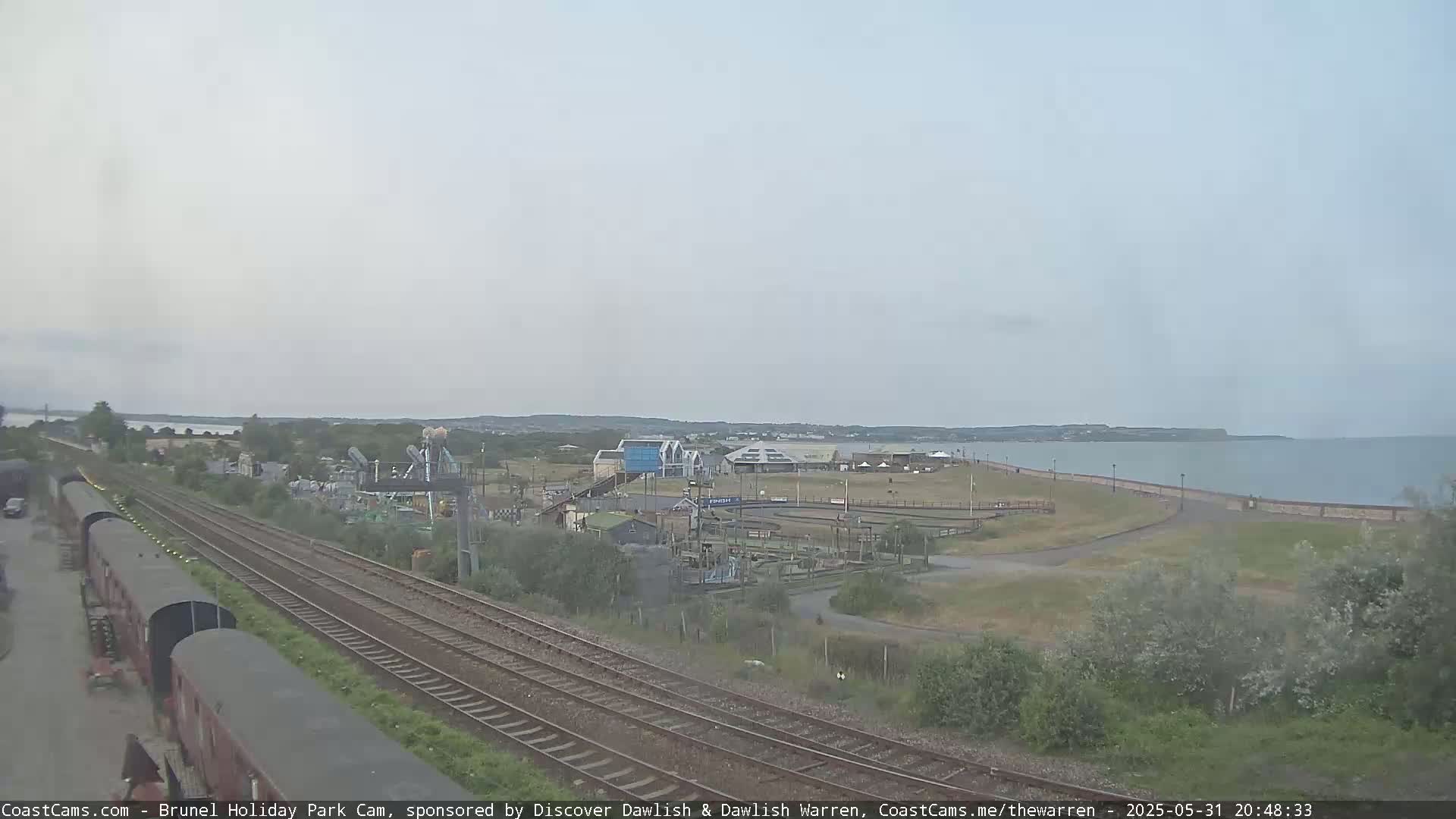 A coastal scene under an overcast sky shows railroad tracks leading toward a beachfront amusement park and calm sea.