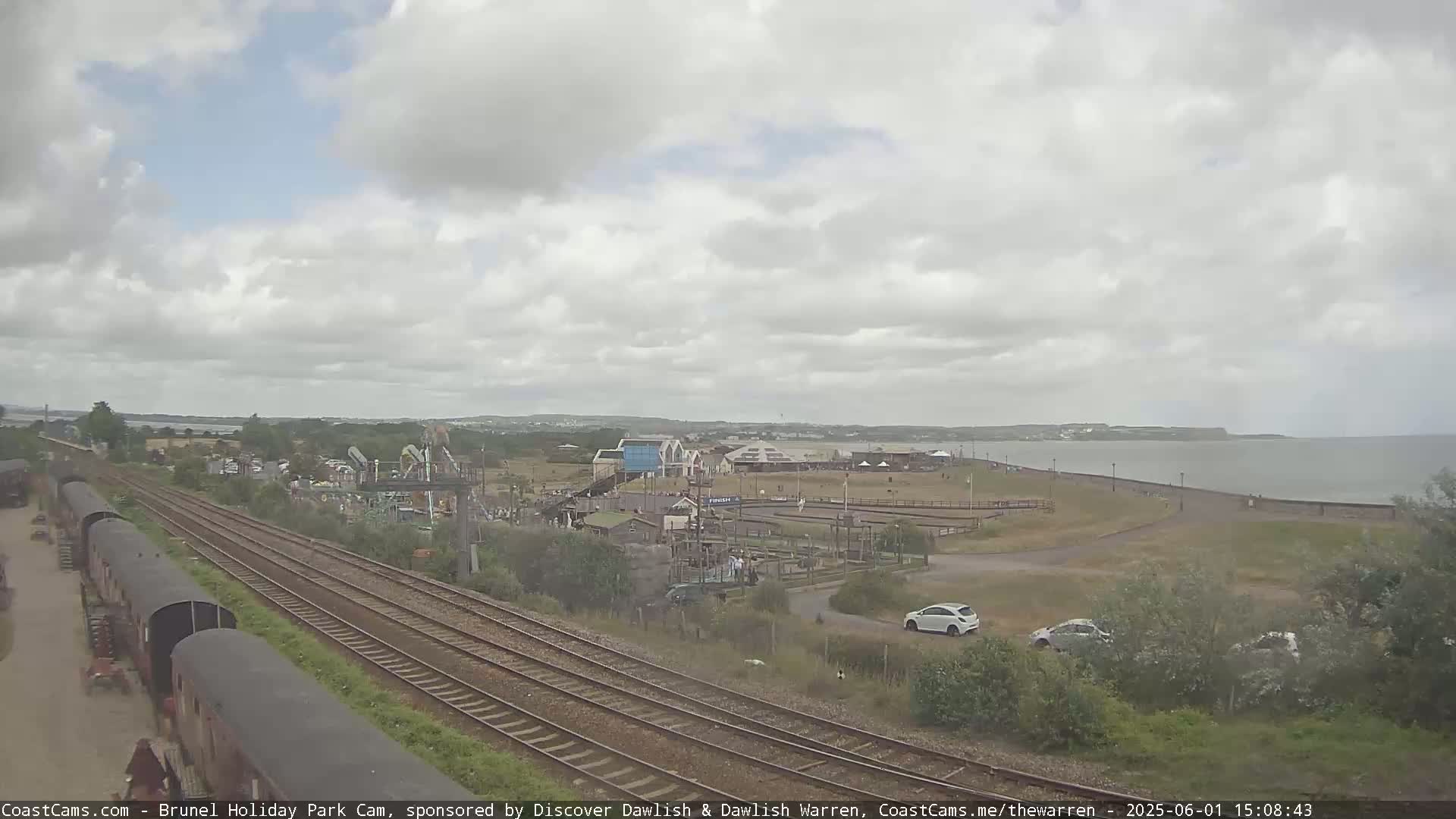 A seaside amusement park is visible beyond a train track on an overcast day, with a calm body of water in the background.