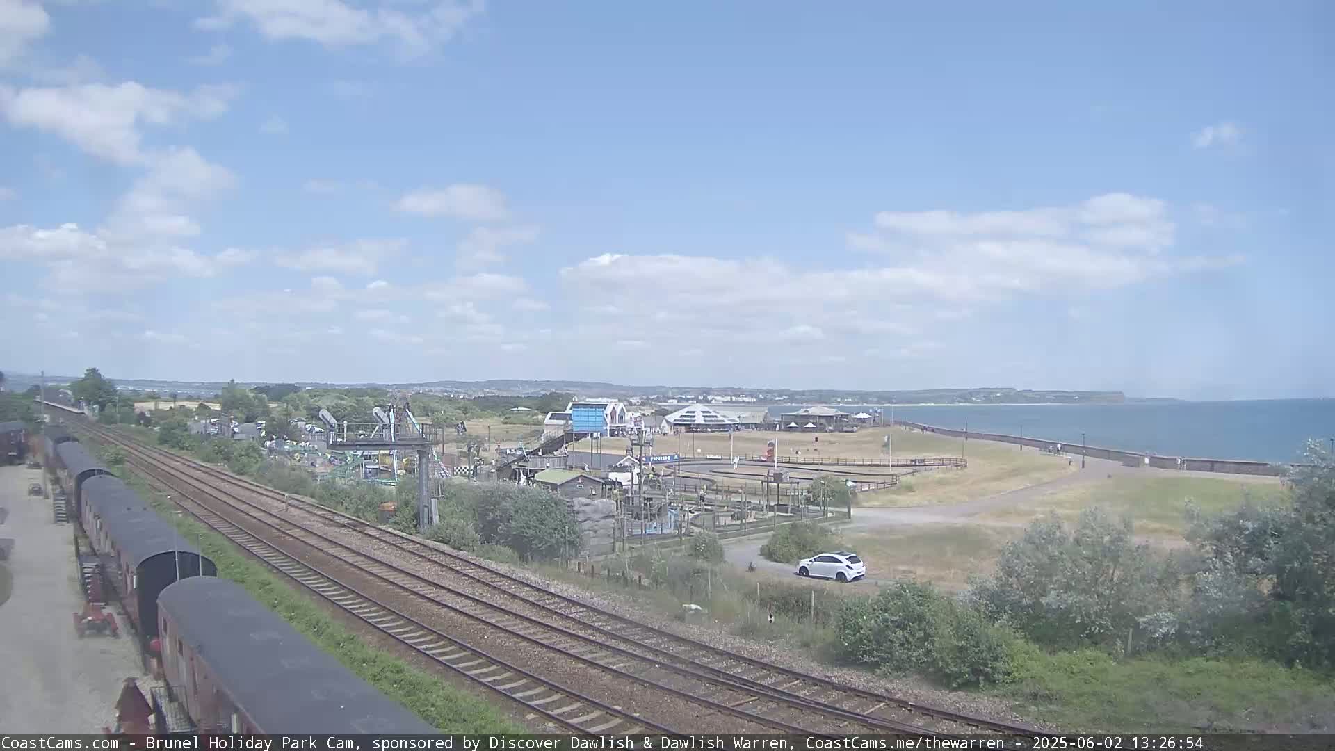A seaside amusement park with a small car park and railway lines is visible under a partly cloudy sky, next to a calm ocean.