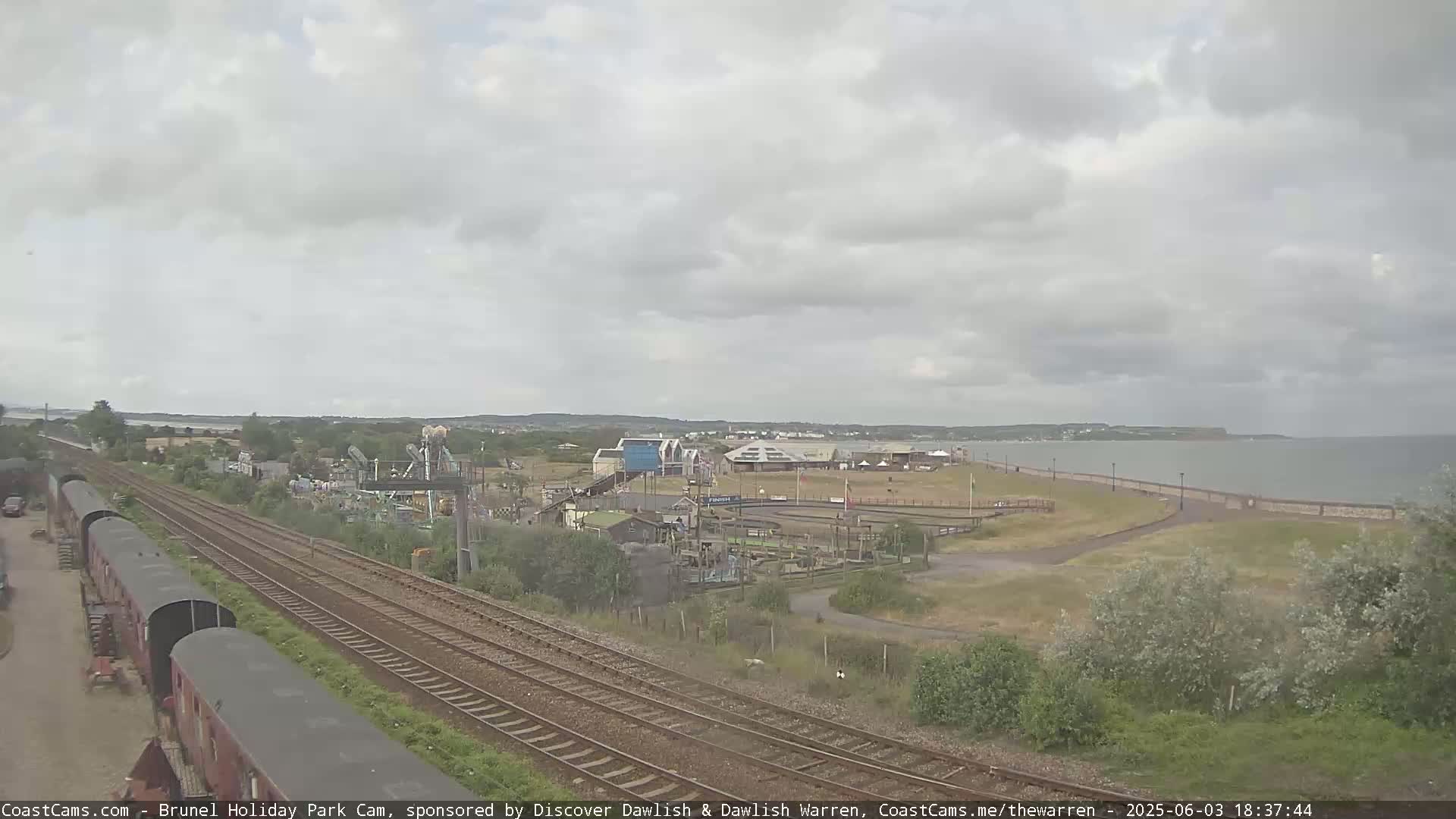 Under an overcast sky, a seaside amusement park is situated next to a body of water and railroad tracks.