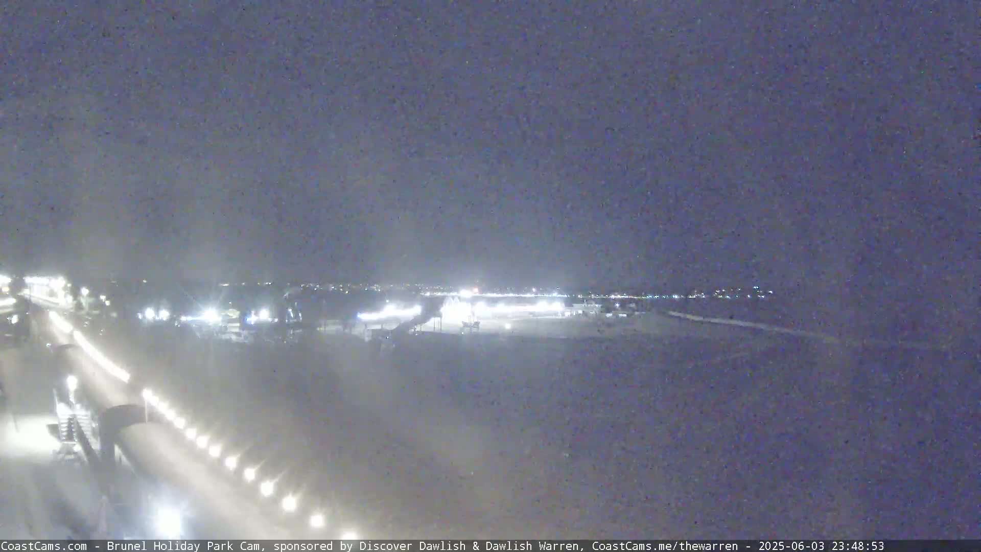 A nighttime, low-light view of a parking lot and roadway with some distant lights and a few structures visible.