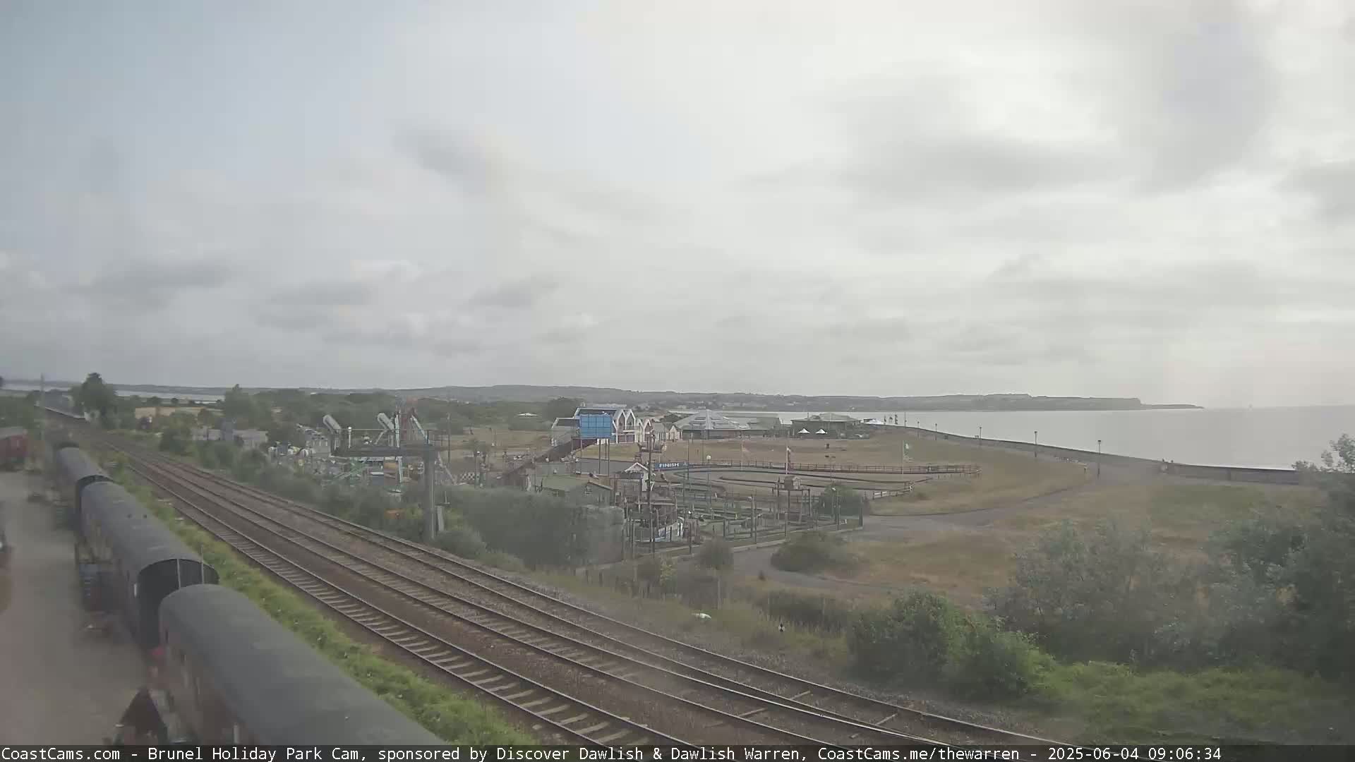 A coastal amusement park with a go-kart track is visible beyond railway tracks under an overcast sky.