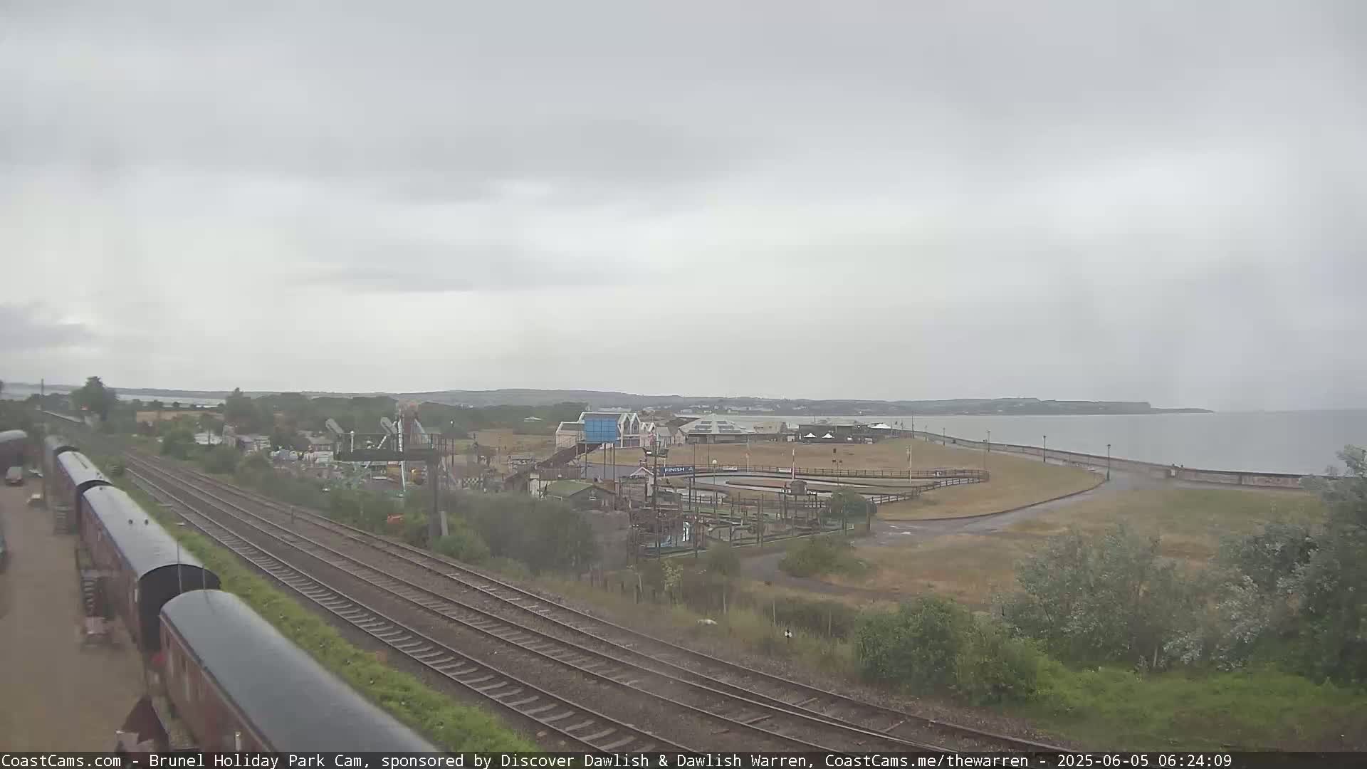 A high-angle, overcast view shows a seaside amusement park near train tracks and a calm body of water.