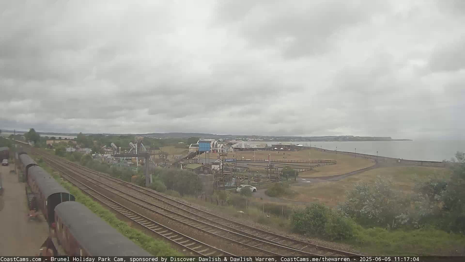 A seaside amusement park is visible beyond railway tracks under an overcast sky.