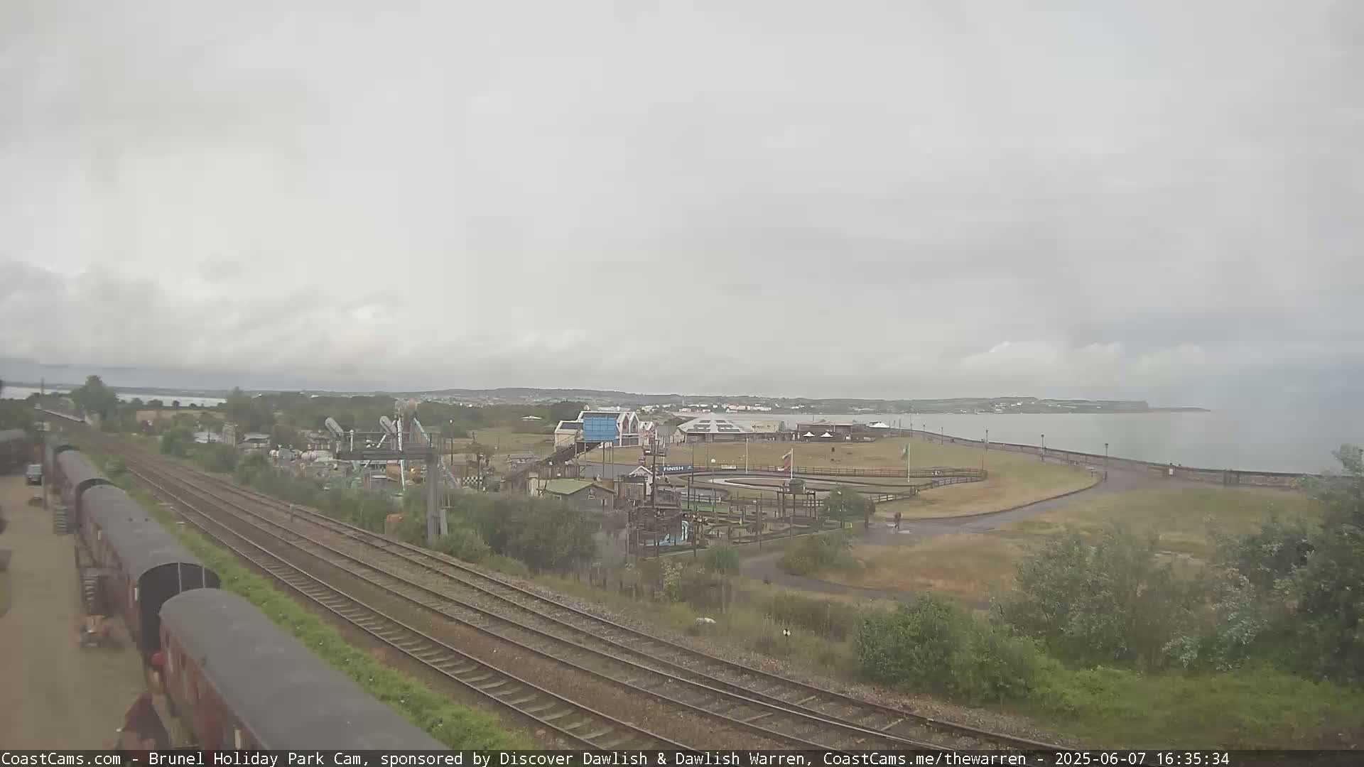 A cloudy day overlooks a seaside amusement park, train tracks, and a calm body of water.