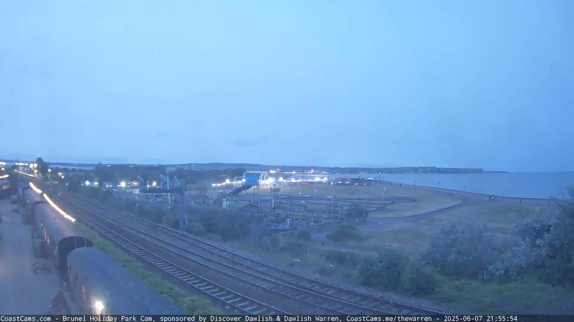 A seaside amusement park at twilight, next to a train track and body of water under a dusky sky.