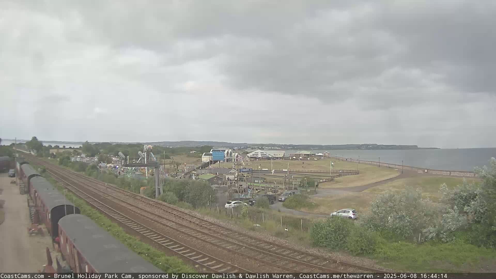 Overcast skies loom over a seaside amusement park, train tracks, and parked cars near a calm body of water.
