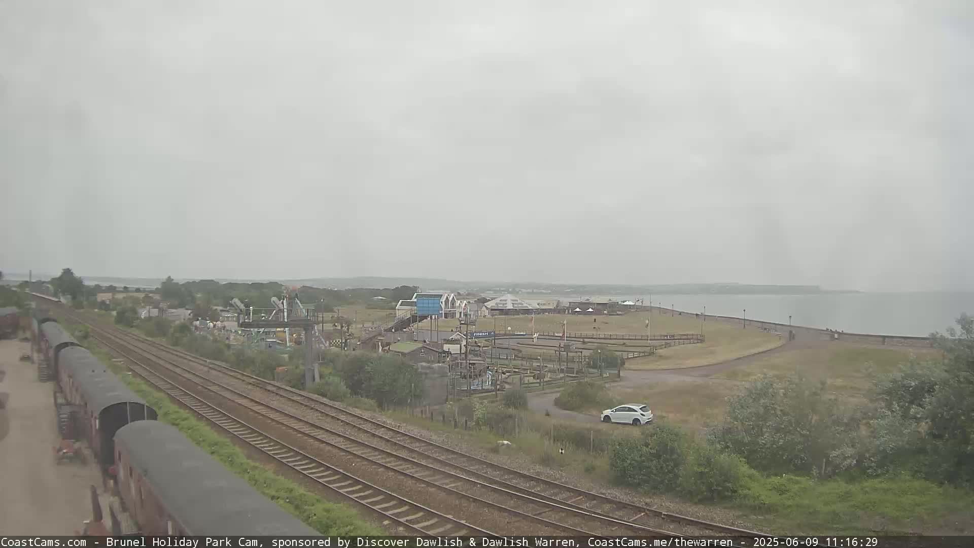 A seaside amusement park is visible next to train tracks, under an overcast sky, with a calm body of water in the background.
