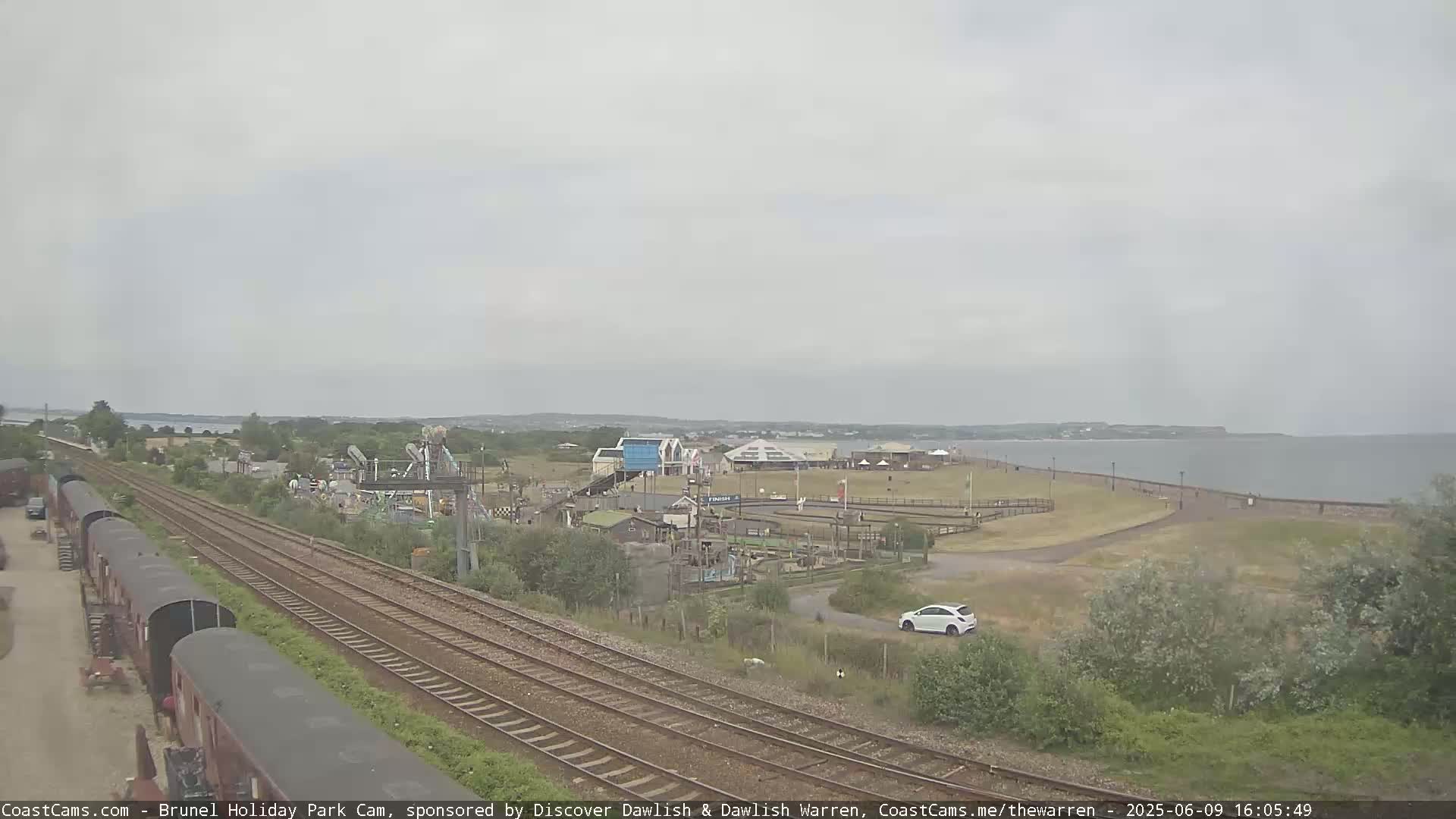 A coastal amusement park with a playground and a small car park is visible next to train tracks under an overcast sky.