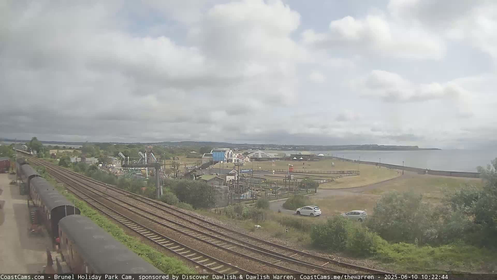 A seaside amusement park with a train track running alongside it is shown under a partly cloudy sky, with calm water visible in the distance.