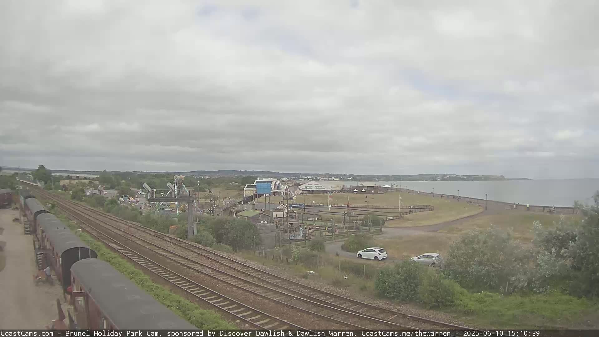 A seaside amusement park with railway tracks and parked cars is visible under an overcast sky.