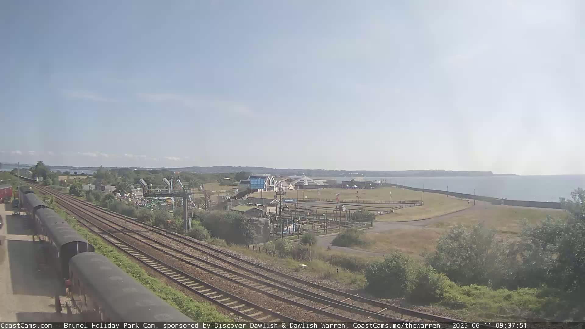A sunny day shows train tracks alongside a seaside holiday park with a calm ocean in the background.