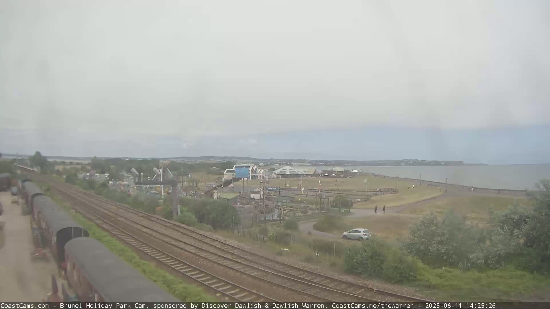 A seaside amusement park with a beach and calm sea is visible beyond train tracks and railway cars under an overcast sky.