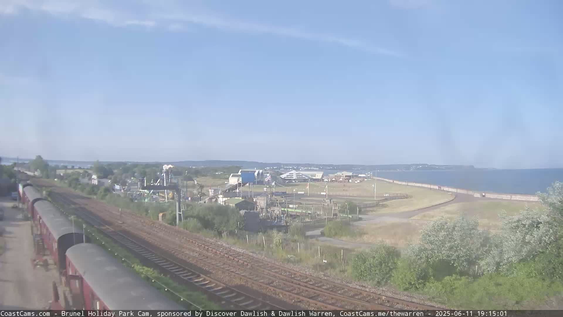 A seaside amusement park is visible beyond railroad tracks on a sunny day, with a calm body of water in the background.