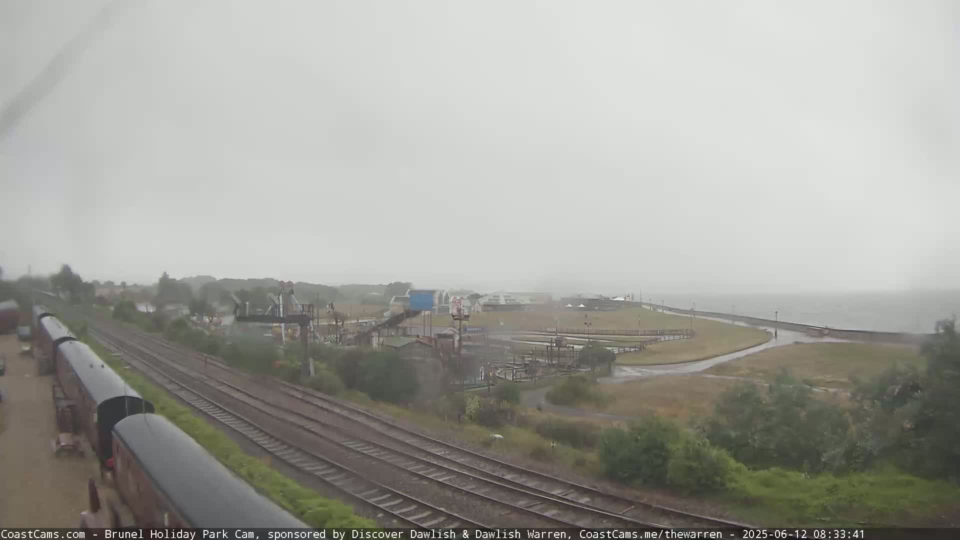 A rainy, overcast day shows a view of railroad tracks next to a seaside amusement park and body of water.