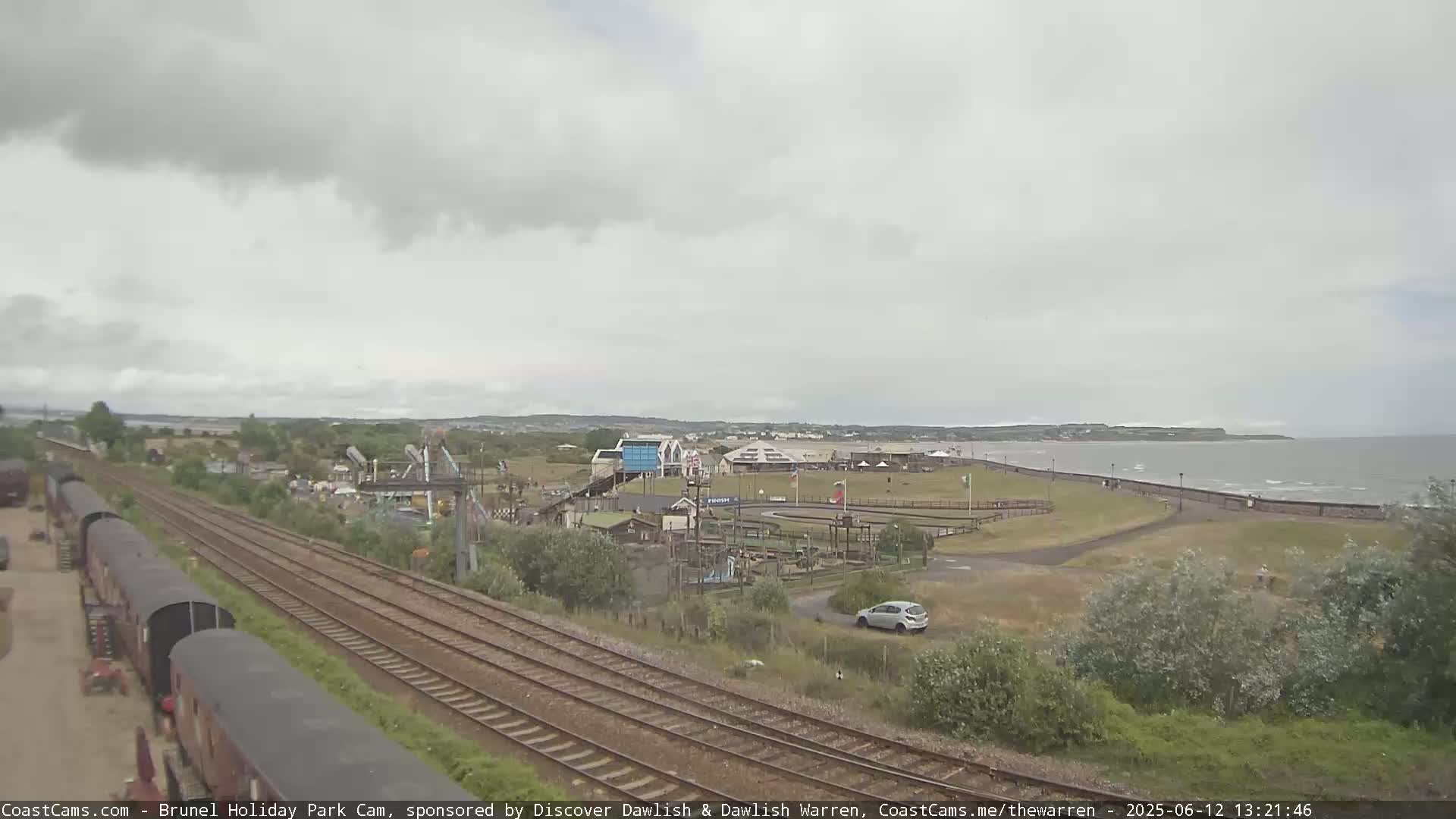 A seaside amusement park with a go-kart track and railway lines is visible under an overcast sky.