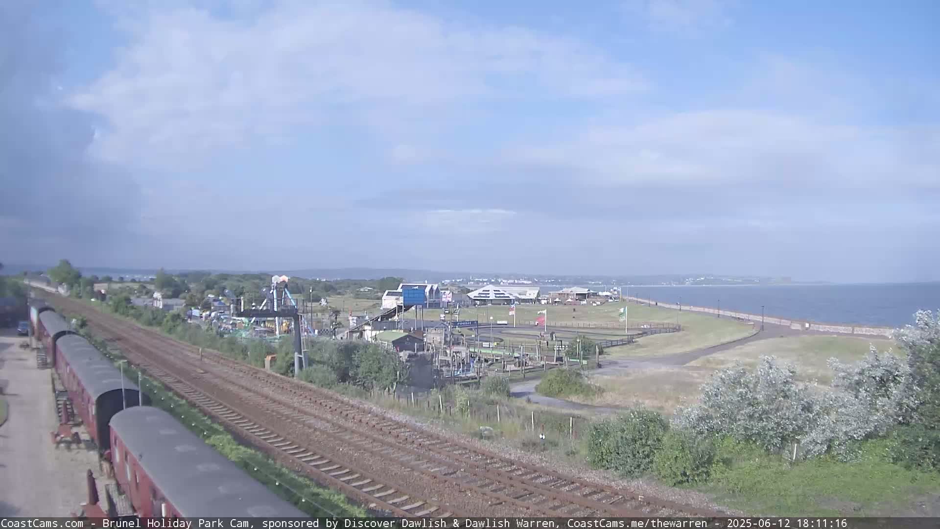 A train track runs alongside a seaside amusement park on a partly cloudy day, with a calm body of water visible in the background.