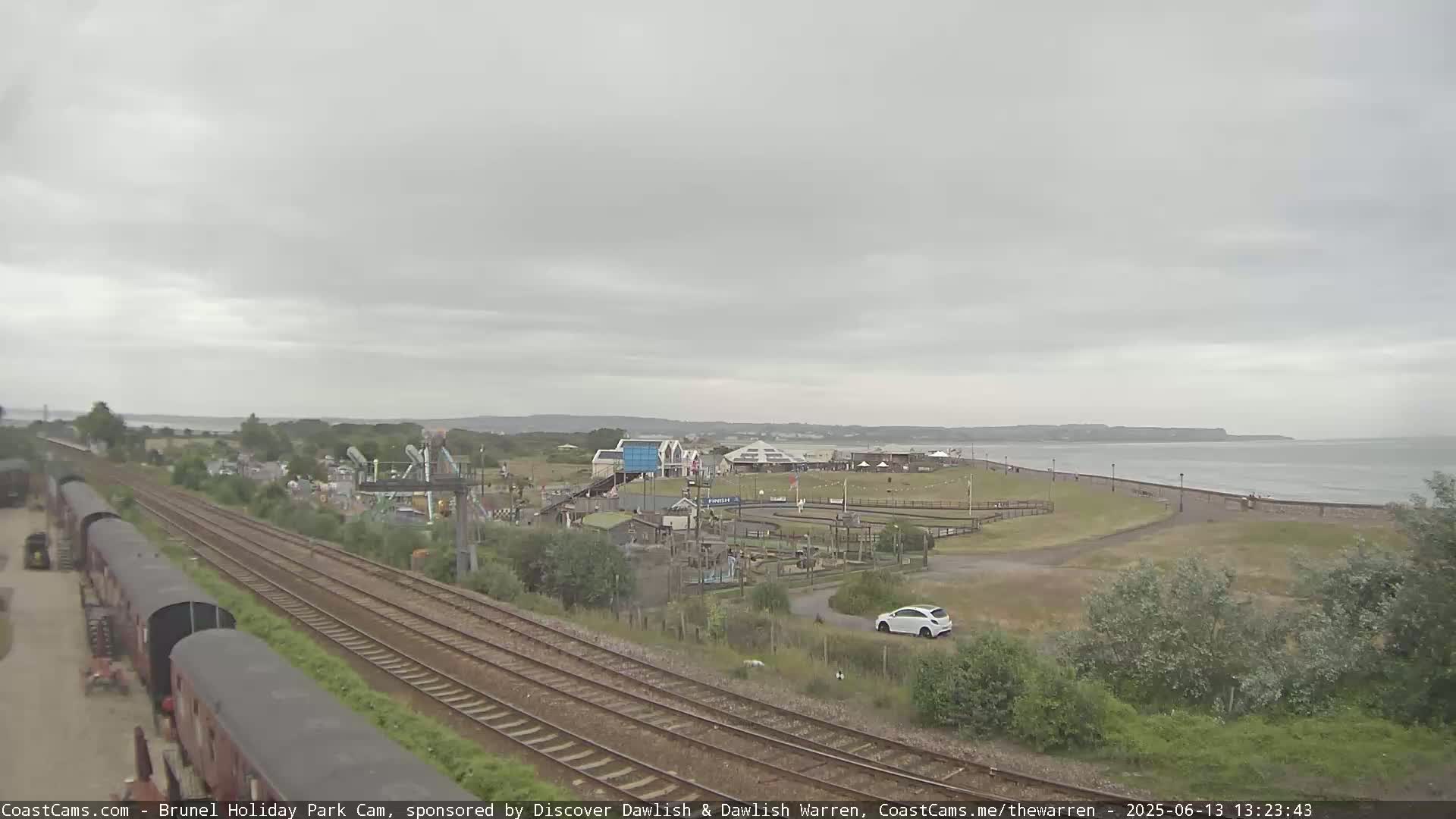 A seaside amusement park is visible beyond a railway track, under an overcast sky and calm sea.